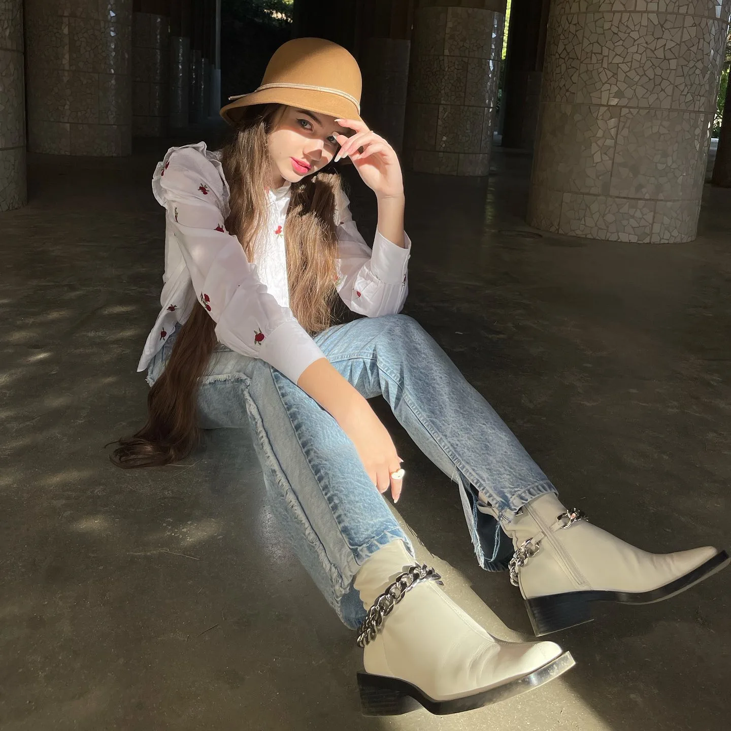 Dana wearing a straw hat, white blouse, and jeans sitting on the floor with chain-adorned boots.