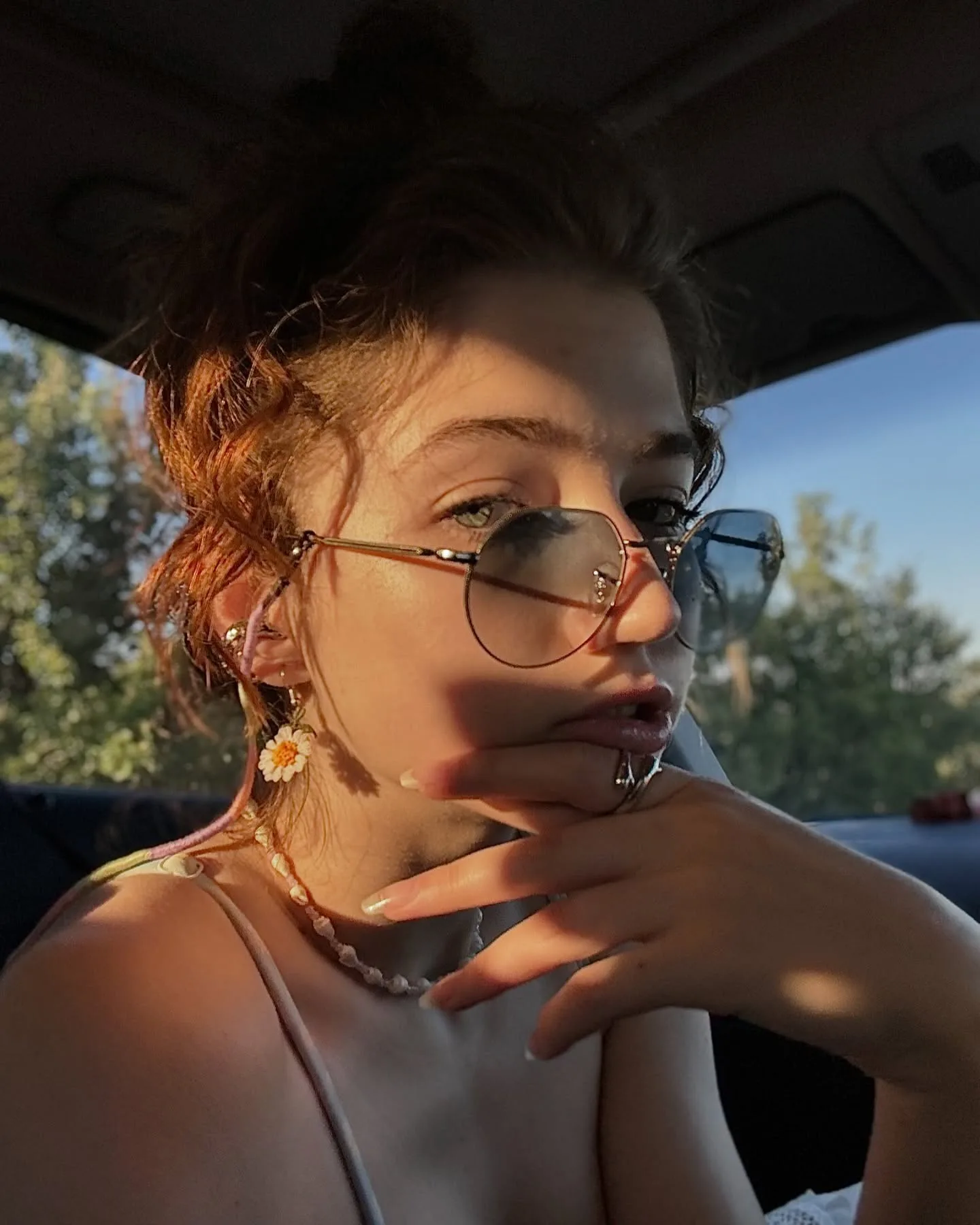 Close-up portrait of a woman with curly hair and glasses sitting in a car, bathed in warm sunlight with her hand near her chin.