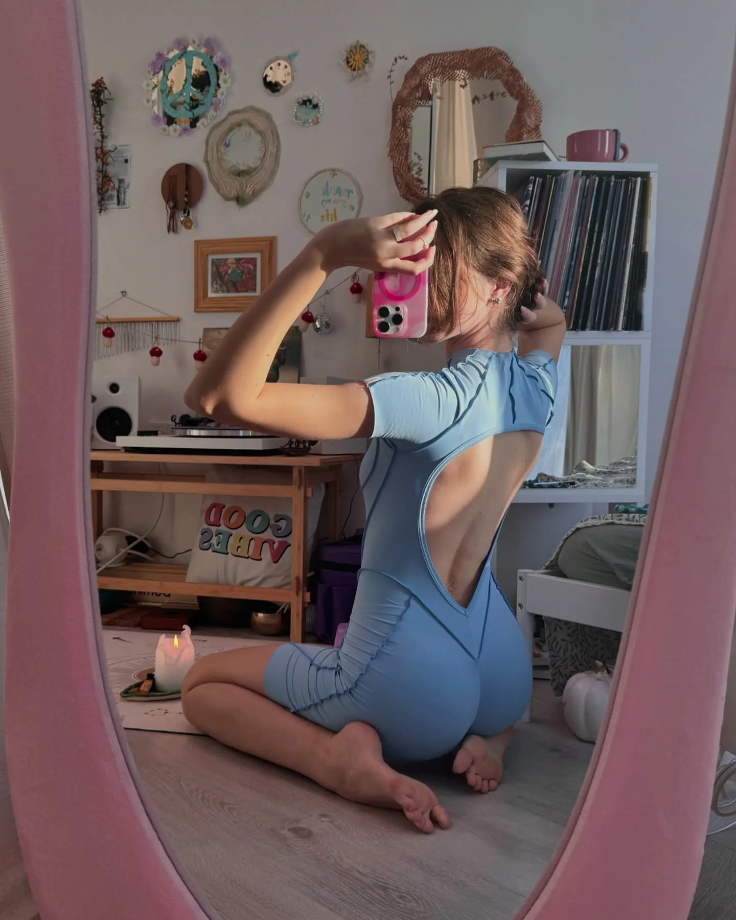 A woman in a blue bodysuit takes a mirror selfie in a pink-framed mirror, kneeling on a wooden floor in a cozy room decorated with wall art and shelves.