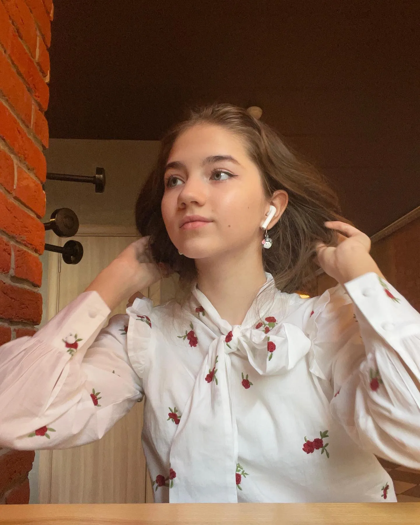 Young woman with brown hair wearing a white floral blouse and white earbuds, adjusting her hair near a brick wall.