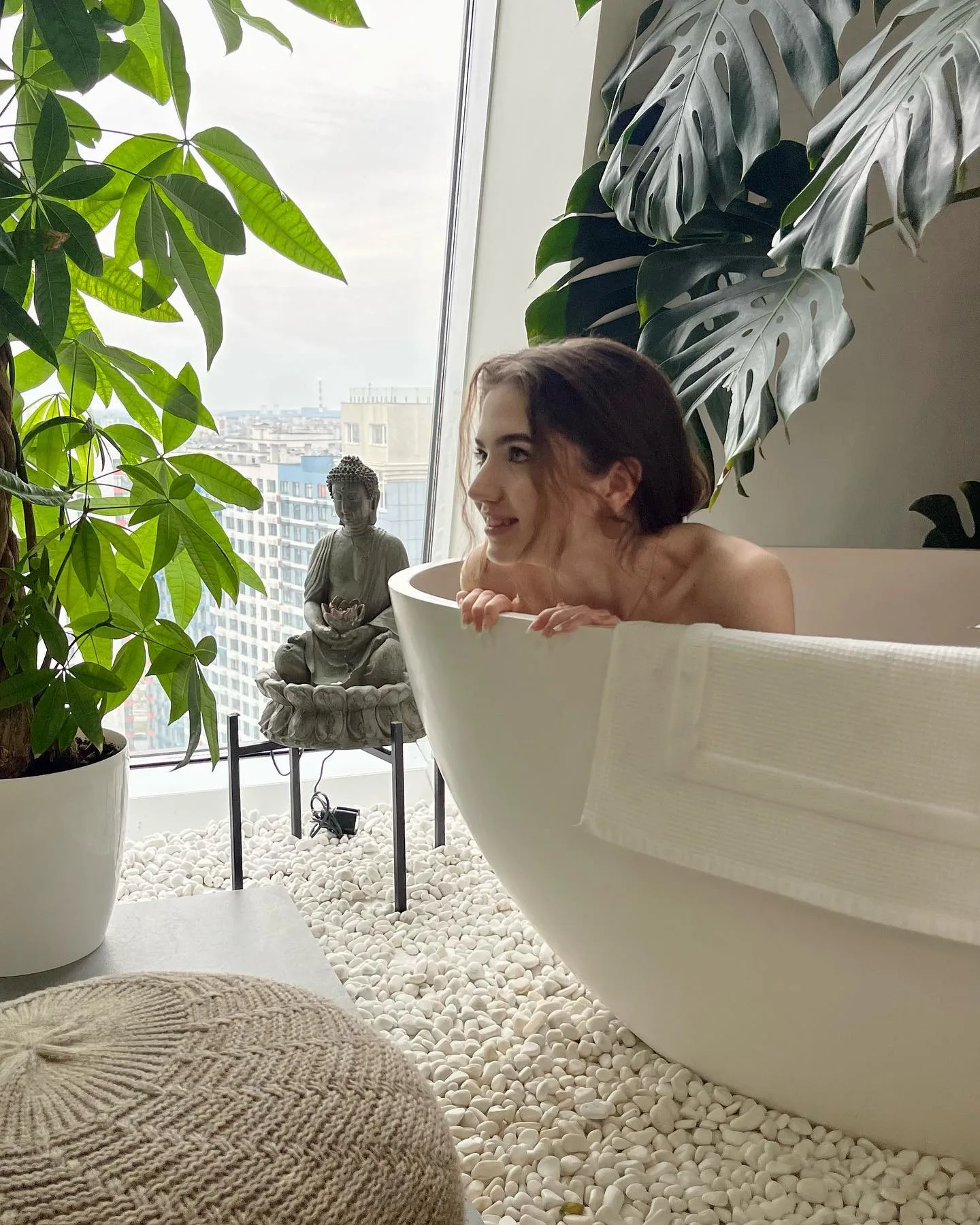 Woman relaxing in a white freestanding bathtub surrounded by lush green plants and white pebbles with a city view.