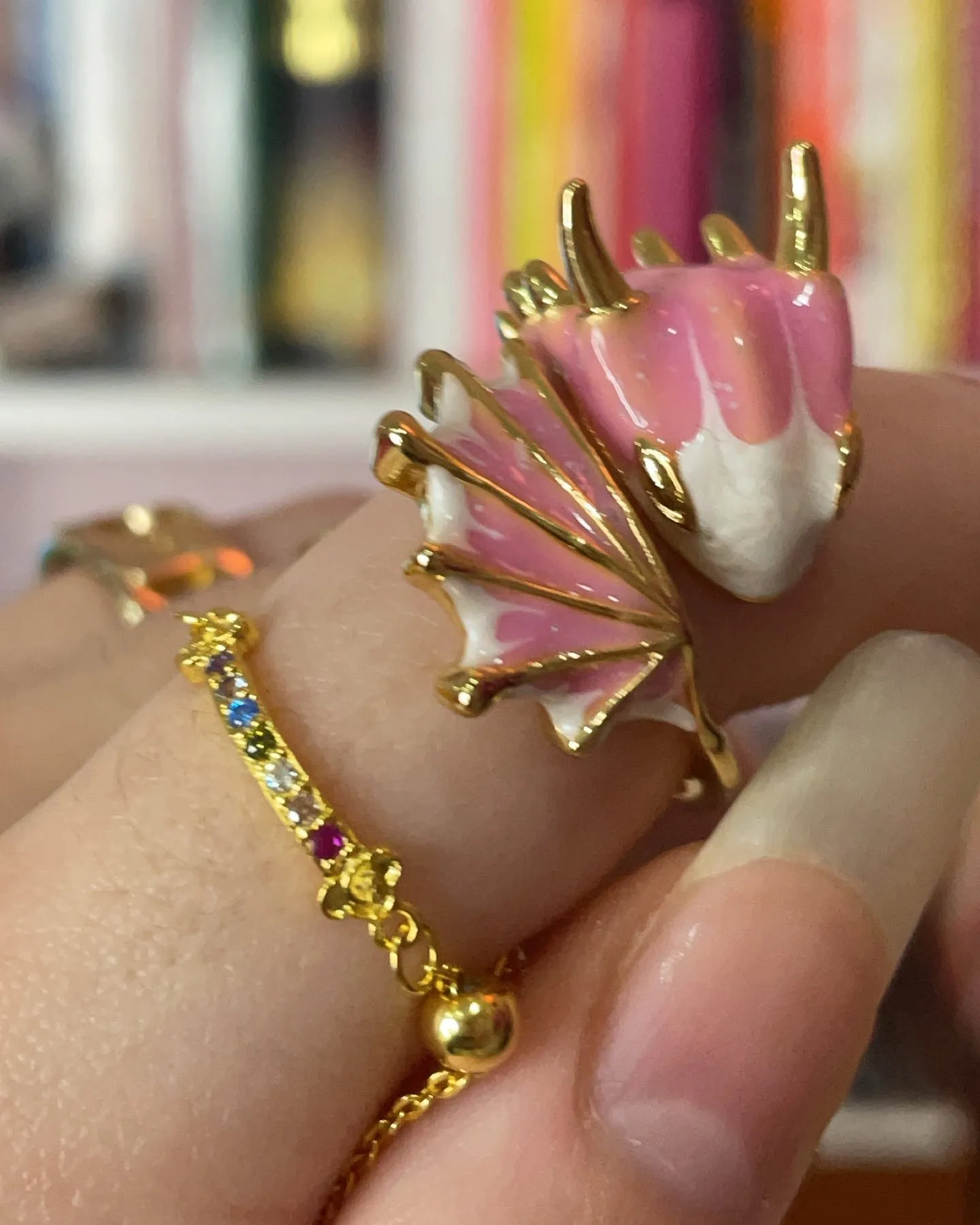 Close-up of a hand wearing a pink and gold enamel dragon head ring and a delicate gold chain ring with colorful gemstones.