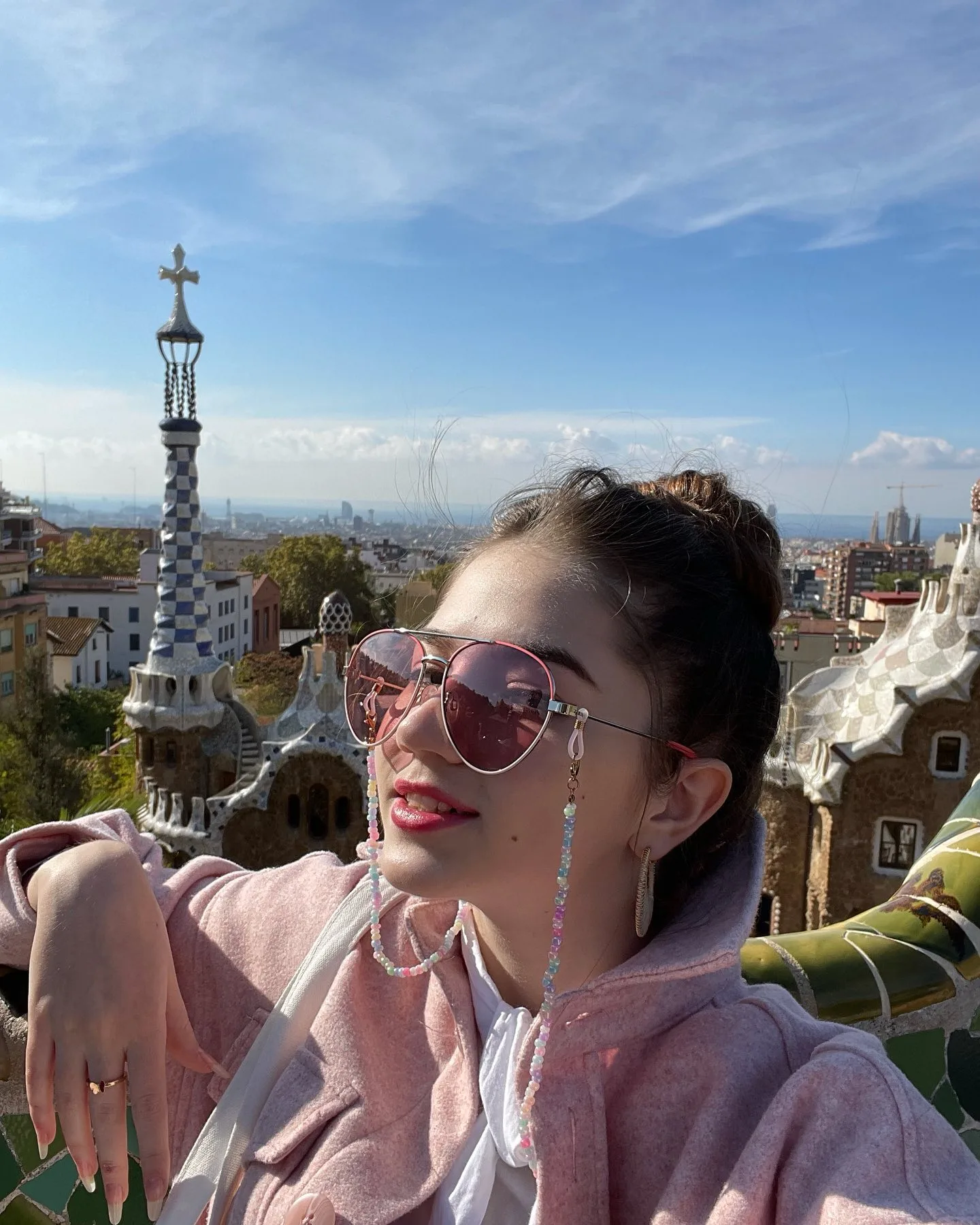 A woman wearing sunglasses and a pink hoodie poses for a selfie with the distinctive mosaic architecture and cross-topped spire of Park Güell in Barcelona visible in the background