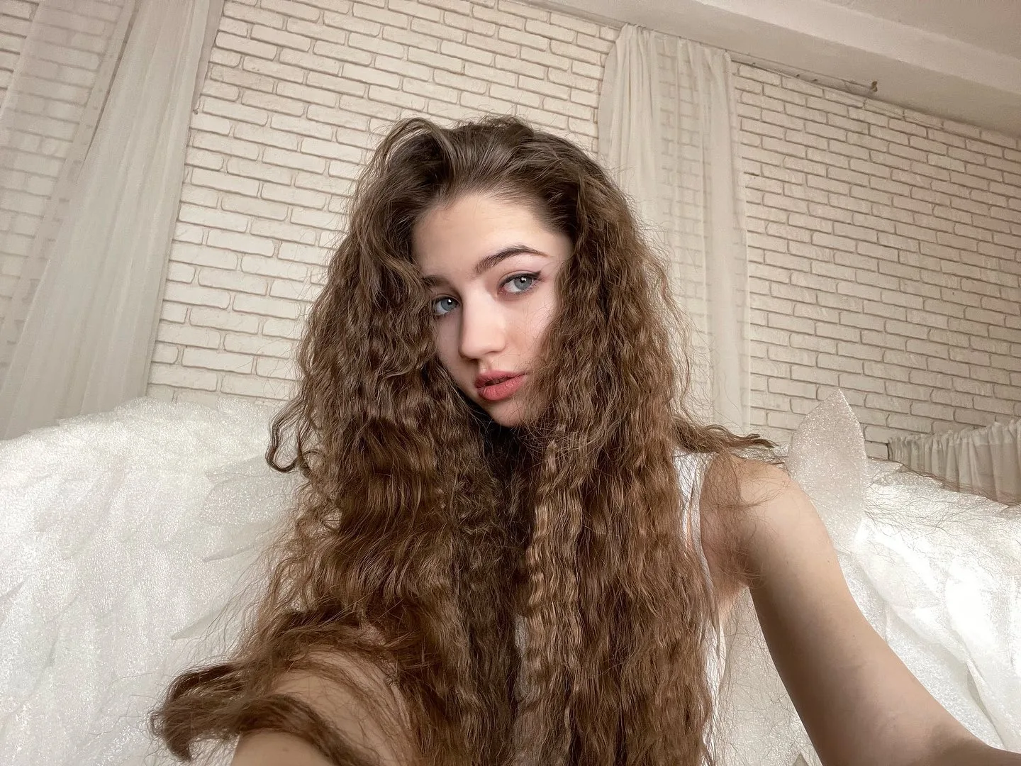 Portrait of Dana with long curly brown hair and blue eyes posing on a white bed with a brick wall background.