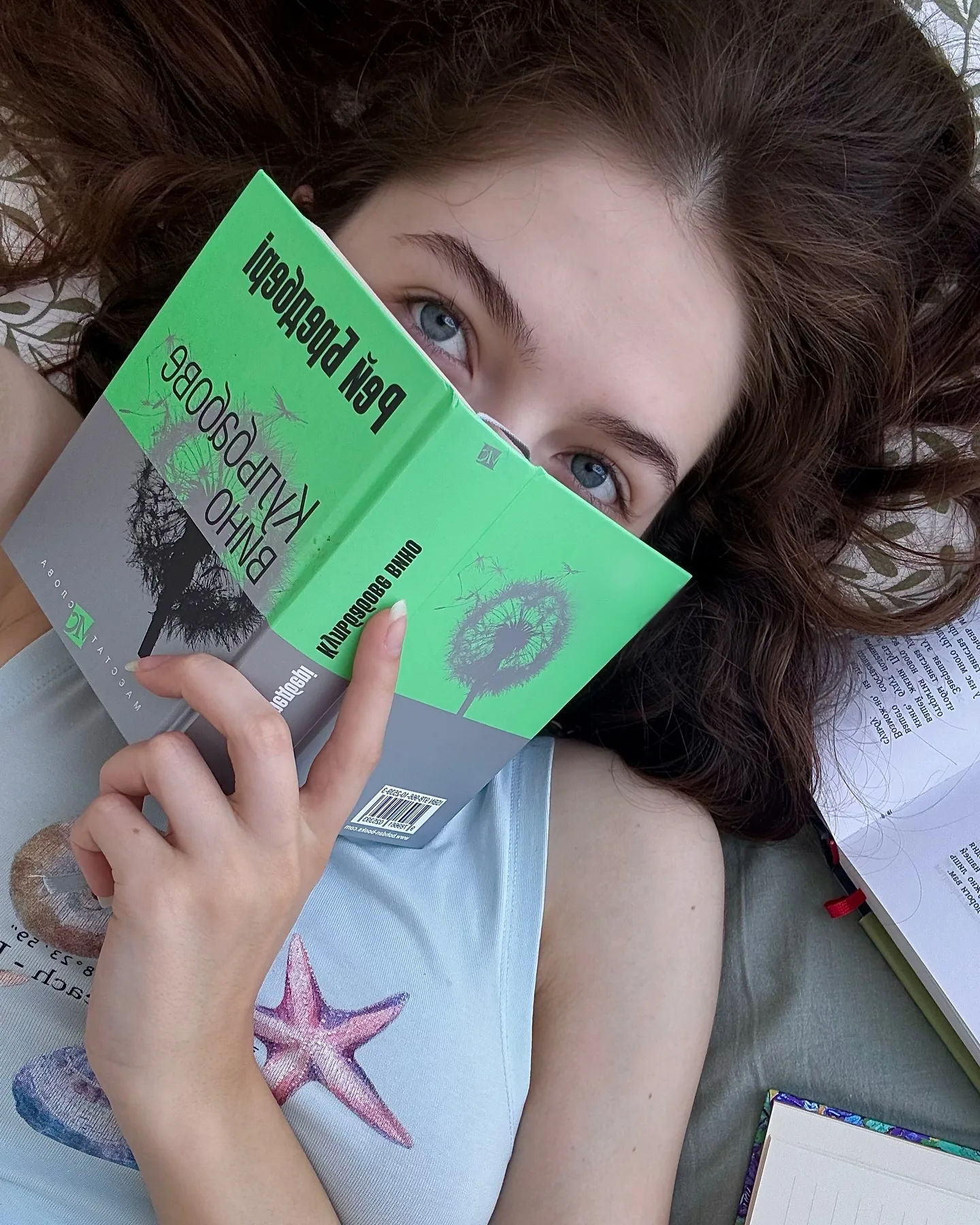 A young woman with brown hair lies on a bed holding a green book titled "The Kite Runner" by Khaled Hosseini over her face, wearing a light blue top with a pink starfish print.