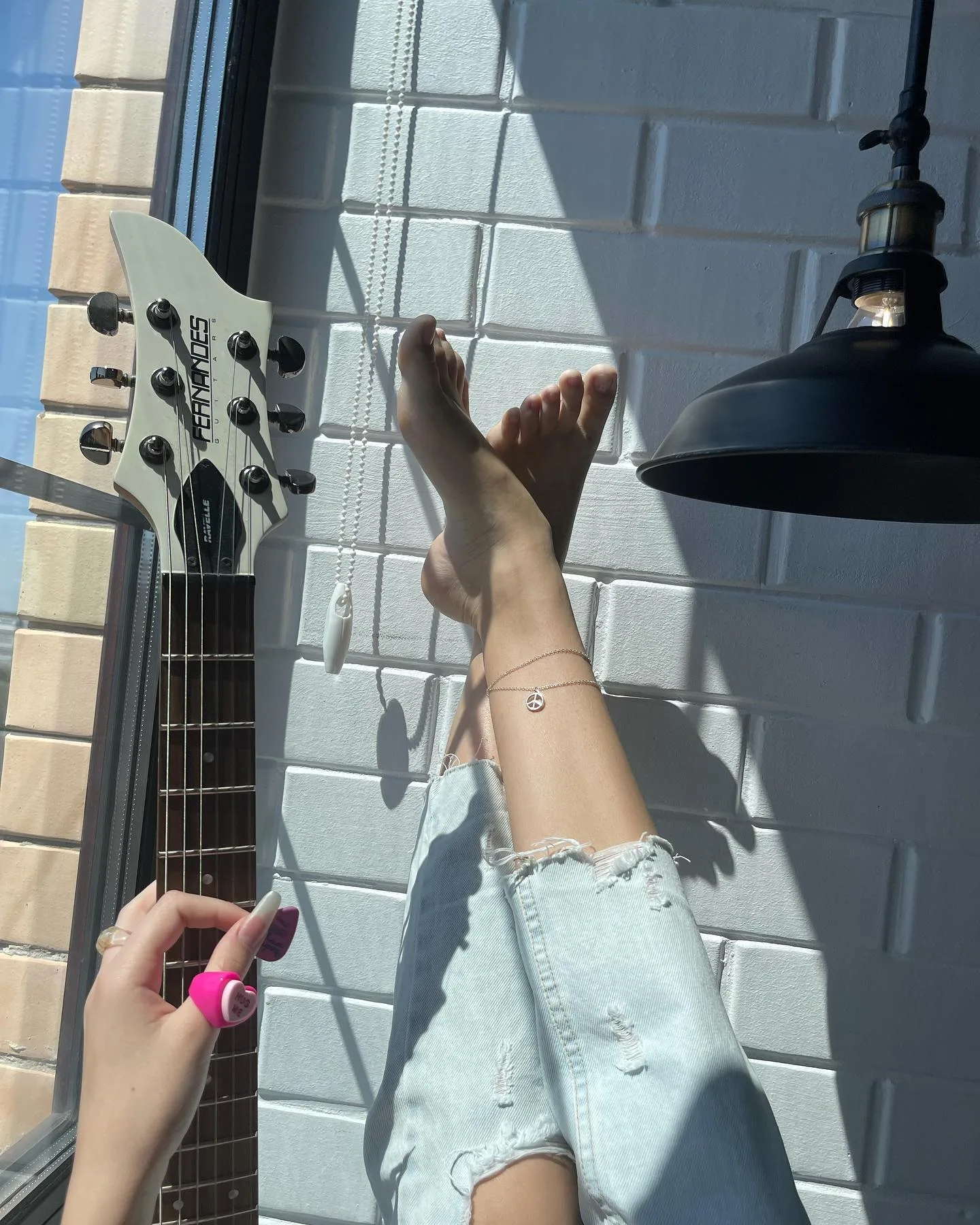 A person painting their toenails pink while relaxing with feet up against a white brick wall next to a guitar headstock.