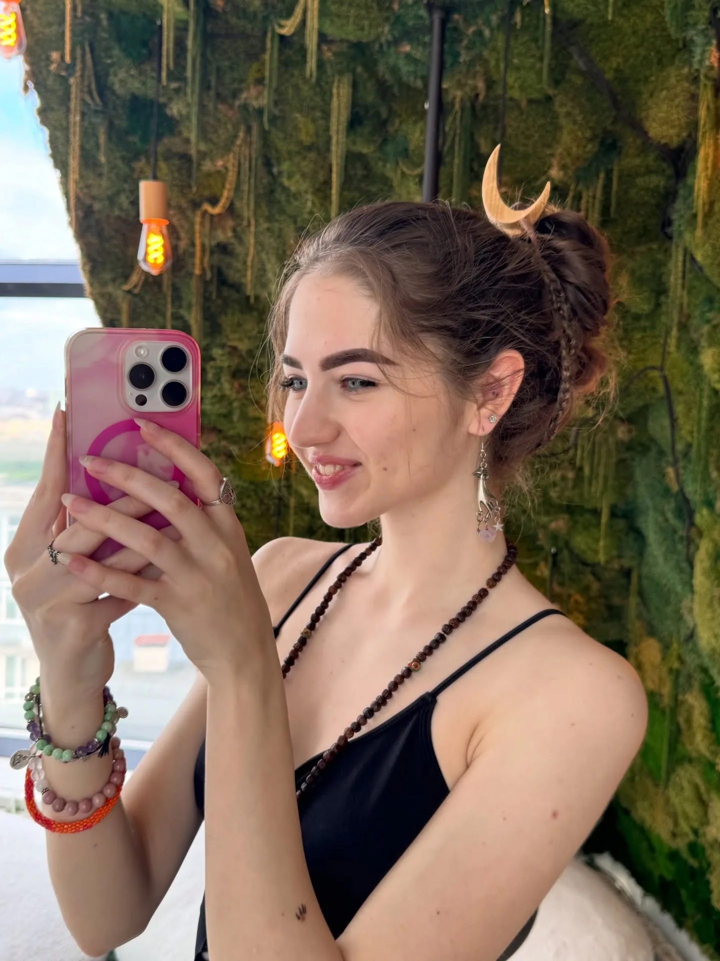 A young woman with brown hair in a bun takes a mirror selfie holding a pink phone, wearing a black top and bracelets against a green moss wall.