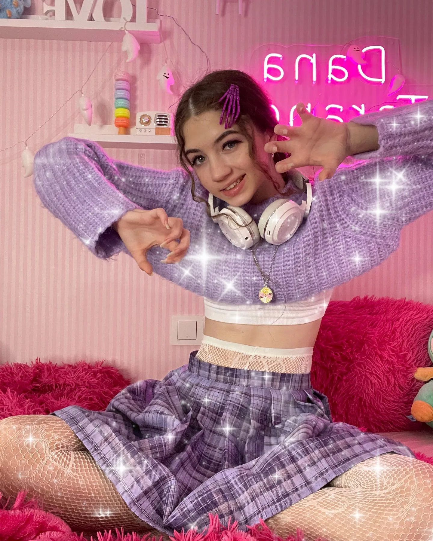 Dana poses in a purple plaid outfit with headphones around her neck, sitting on a pink fluffy rug in a room with pink walls and a neon sign.