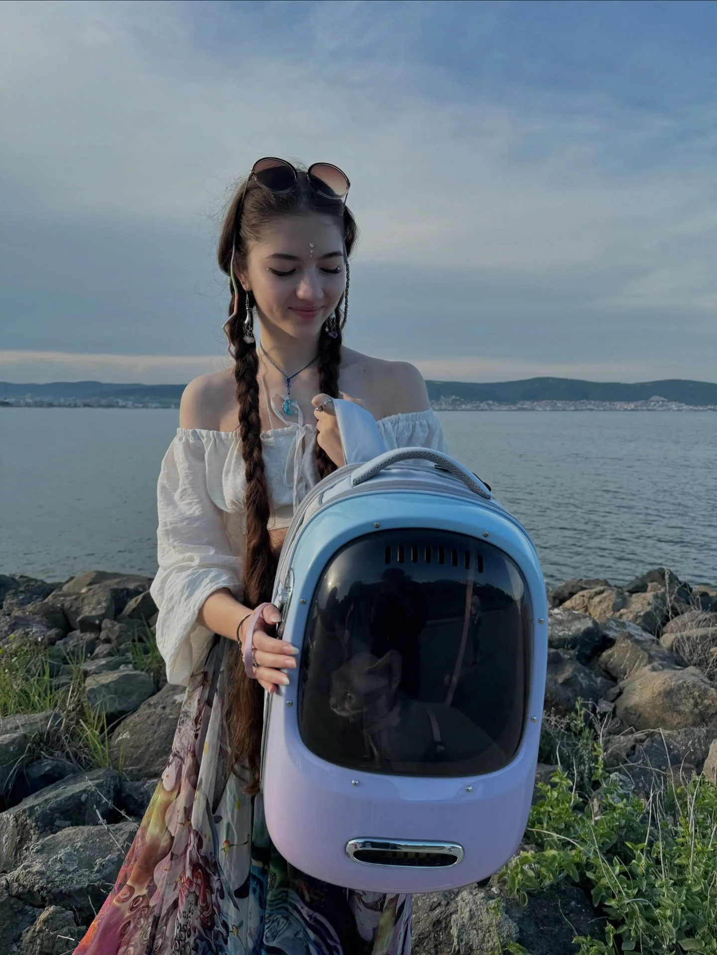 A woman with braided hair stands on rocky terrain near a lake holding a futuristic blue and white pet carrier backpack with a cat inside.