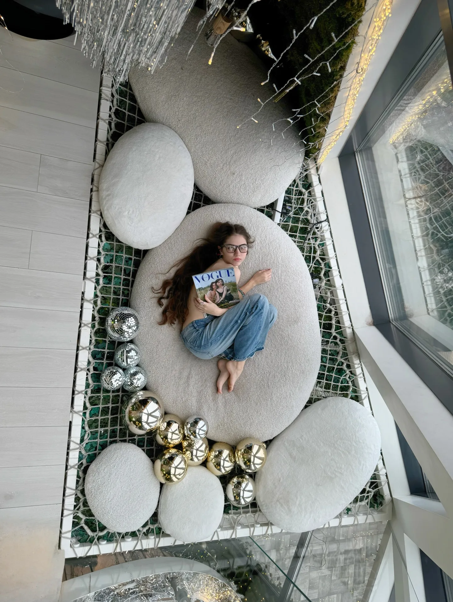 A woman relaxing on a large white cushion surrounded by silver and gold Christmas ornaments in a modern indoor space.
