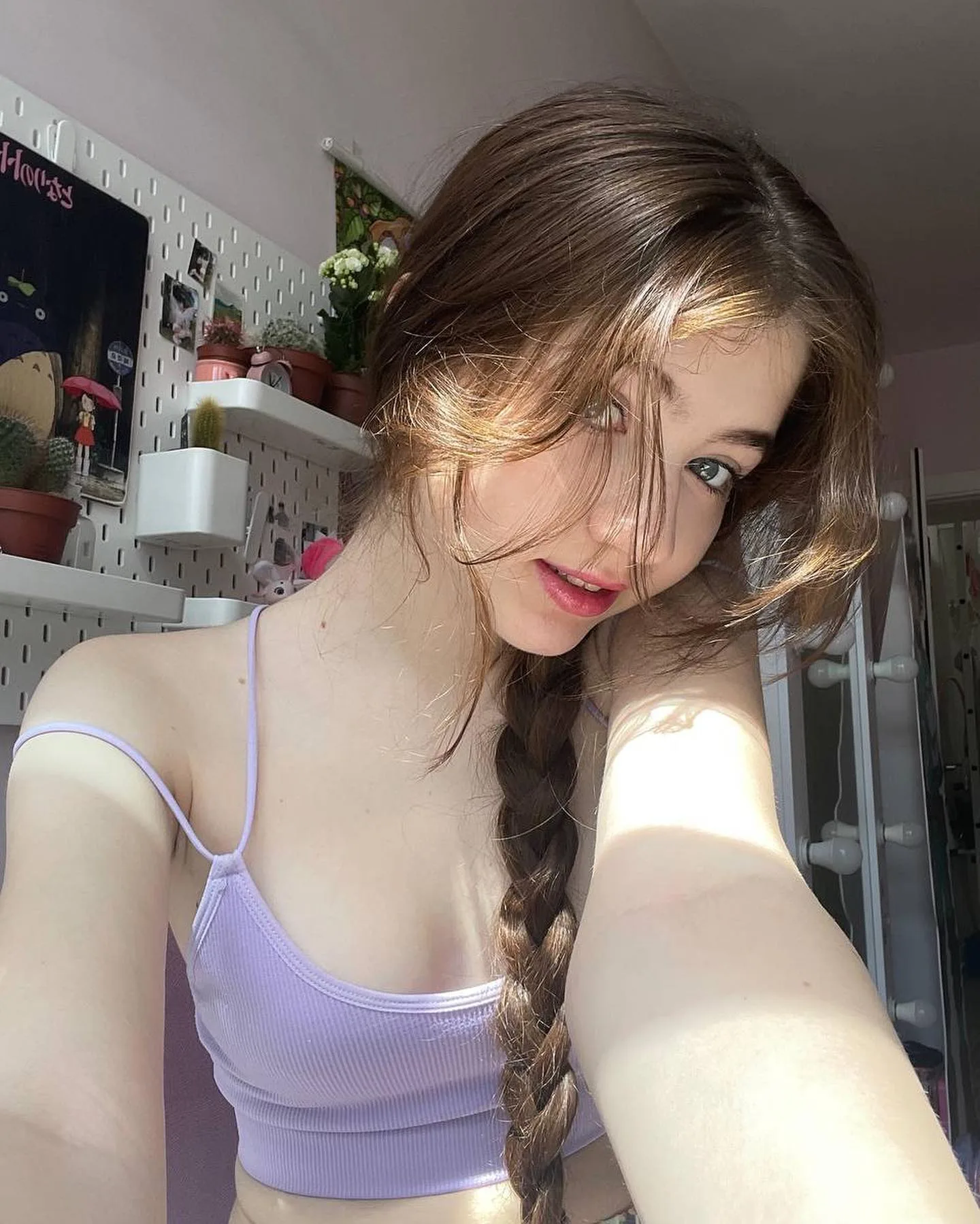 Young woman with long brown hair in a side braid wearing a lavender crop top, posing in a sunlit room with a pegboard wall and plants in the background.