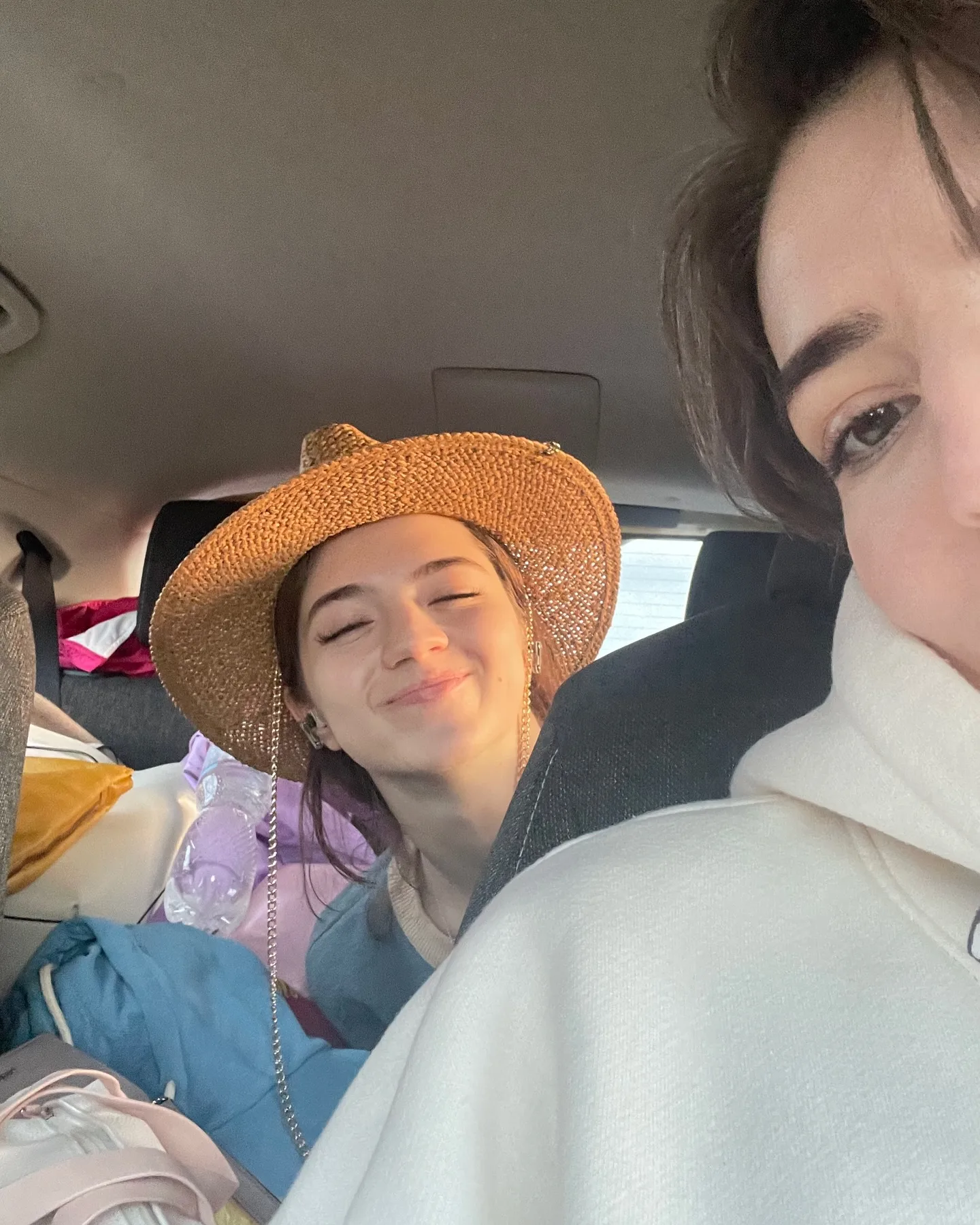 Two women sitting in a car, one wearing a straw hat and smiling with eyes closed, the other in a white hoodie taking a selfie.