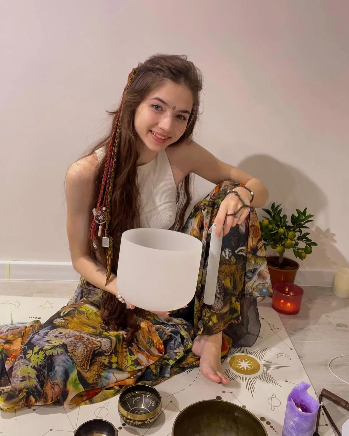 Dana sitting on the floor holding a white singing bowl surrounded by other bowls and a candle.