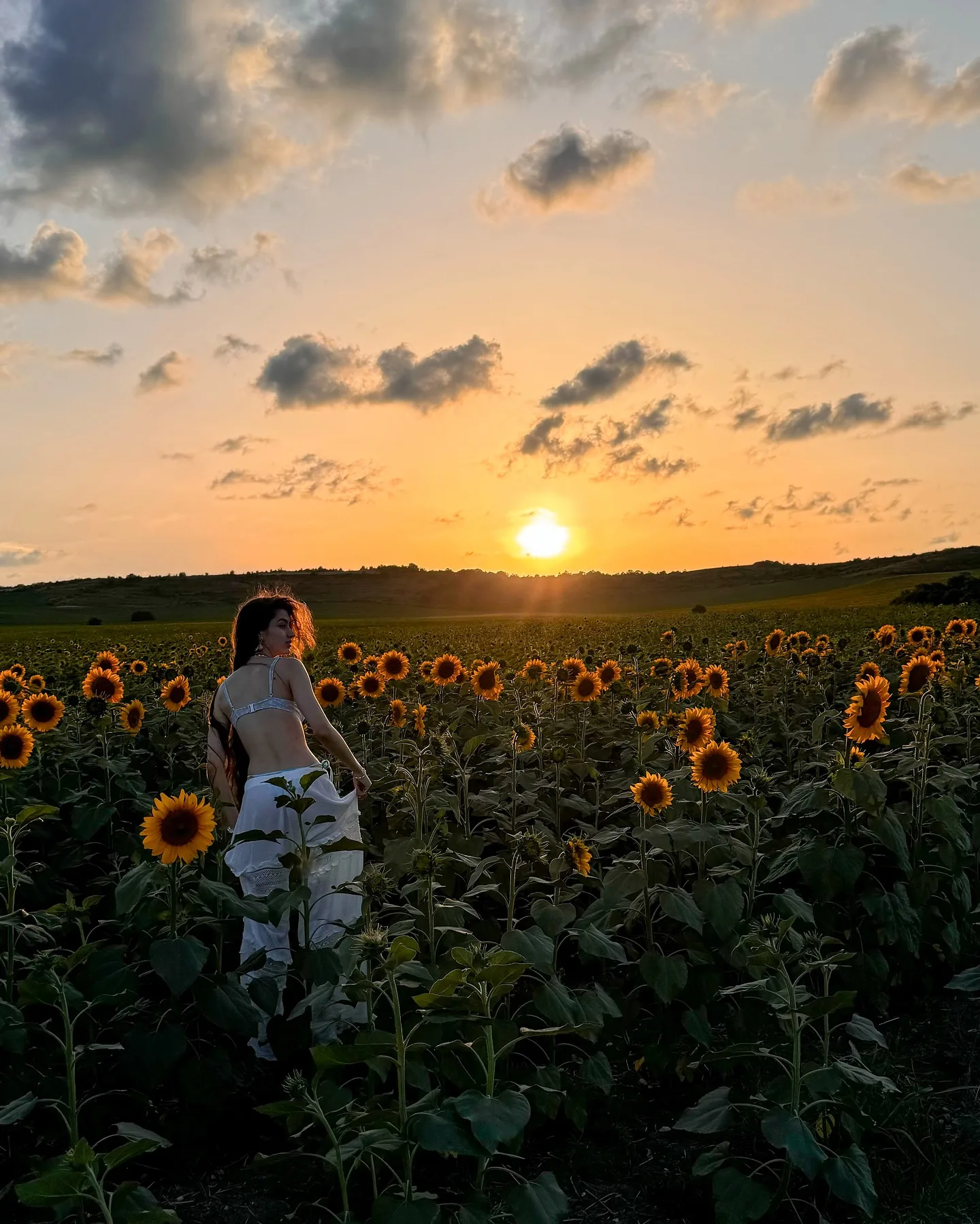 A woman stands in a sunflower field during a golden sunset, with the sun setting on the horizon and scattered clouds in the sky.