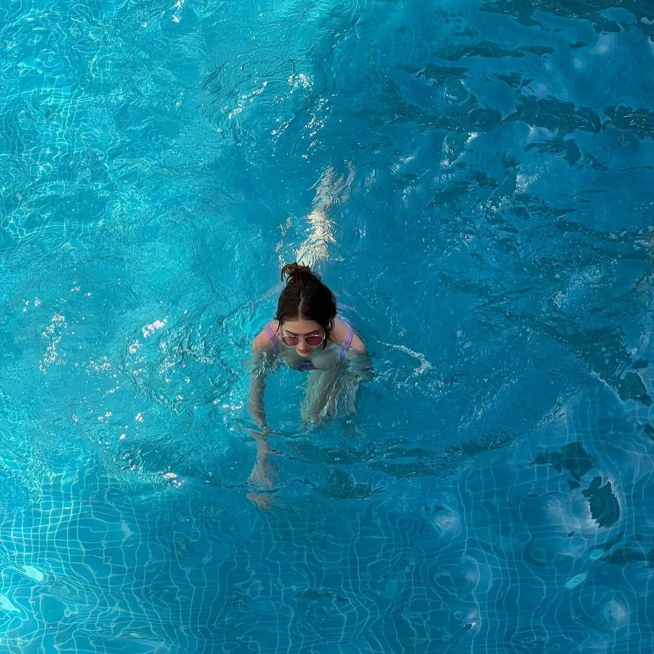 A woman wearing goggles swims underwater in clear blue pool water, viewed from above.