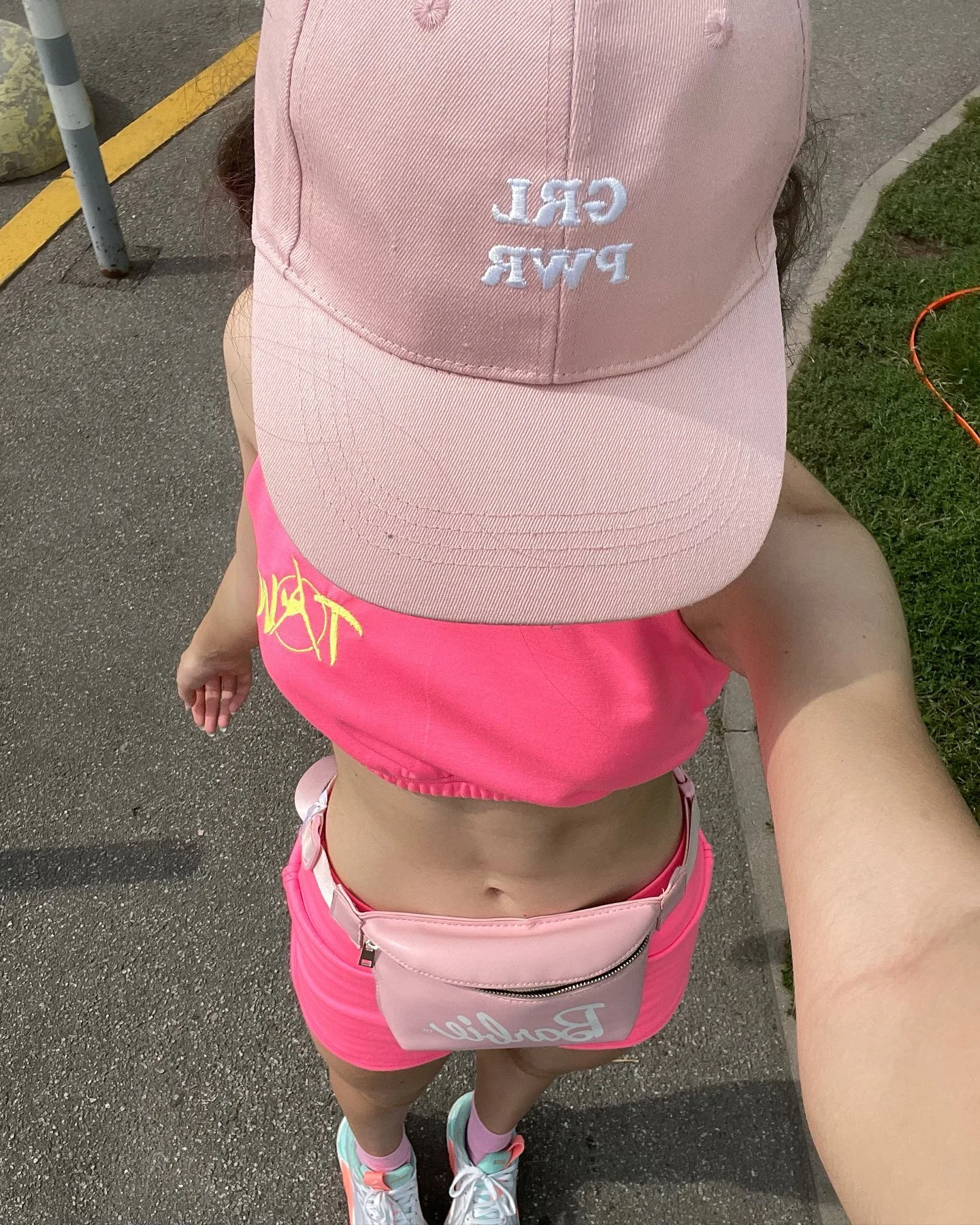 A young girl wearing a pink cap with "GRL PWR" text, a pink crop top, and matching shorts stands on a paved path.