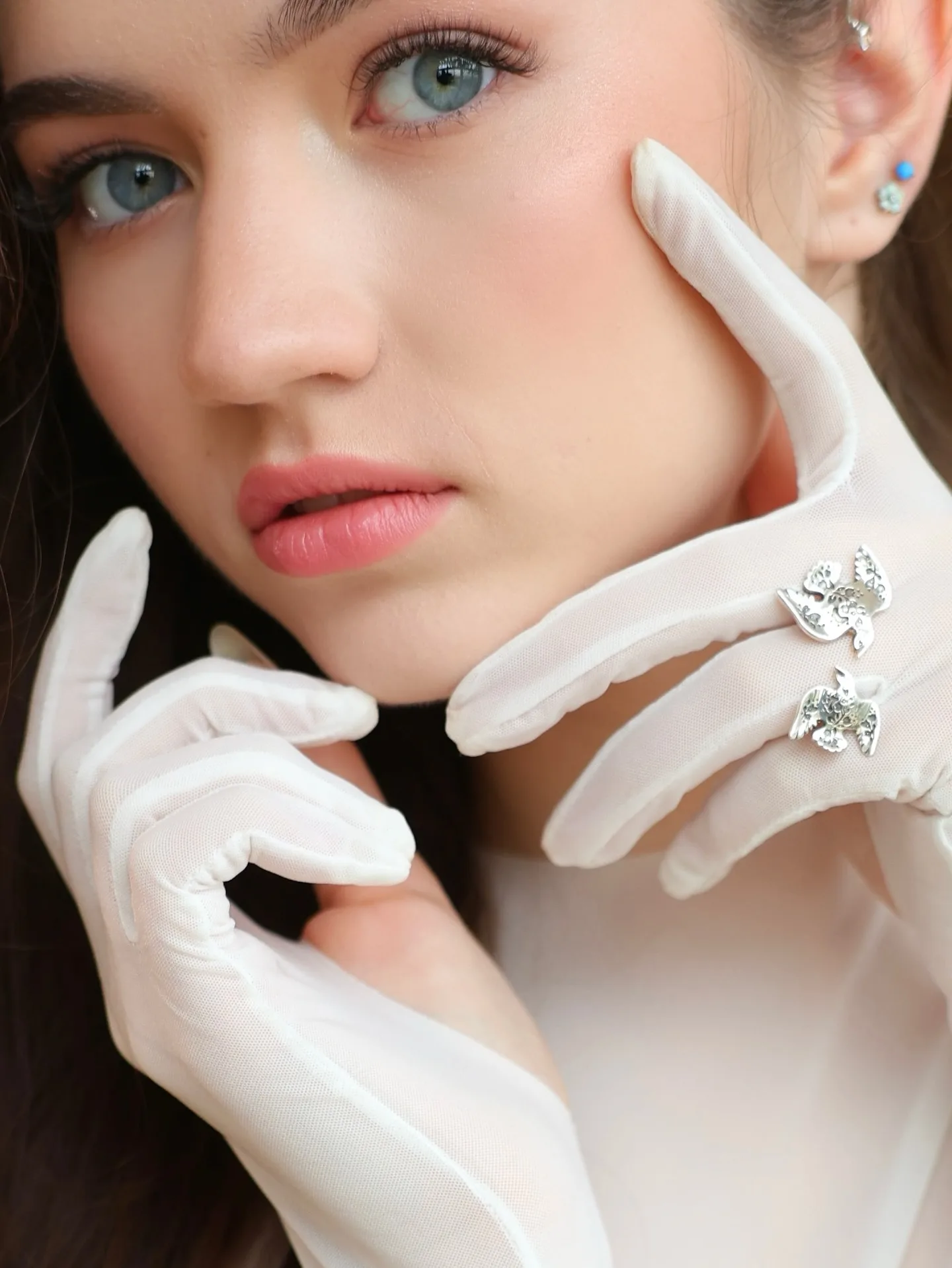 Close-up portrait of a woman with blue eyes wearing white gloves and butterfly rings, posing with hands near her face.