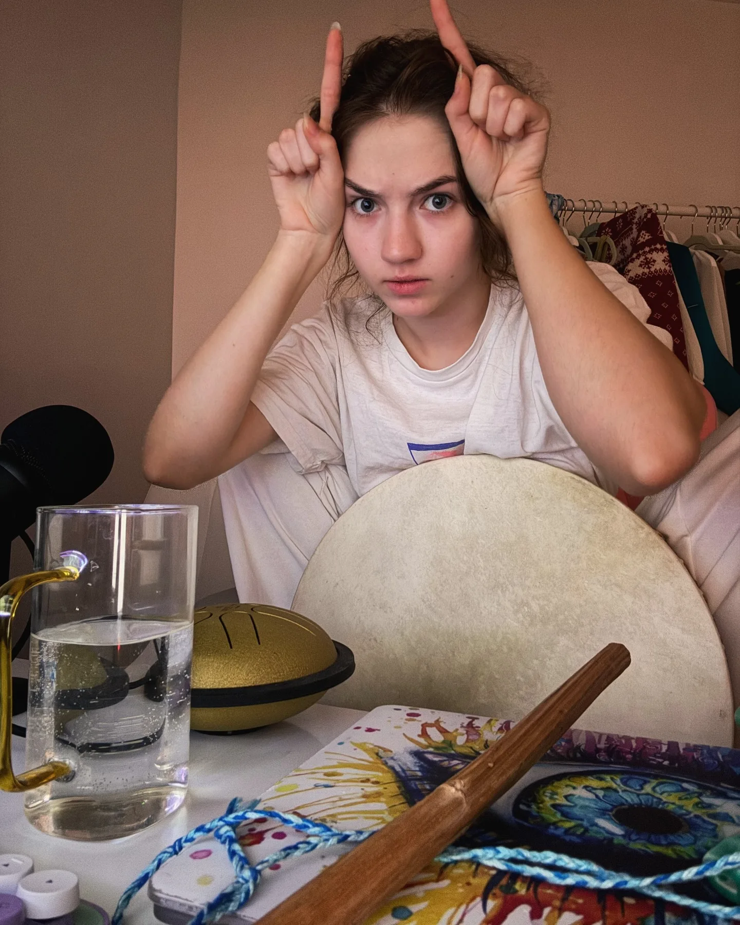 A young woman with brown hair makes devil horns with both hands while sitting at a table with a glass of water, a handpan, a frame drum, and a wooden stick.