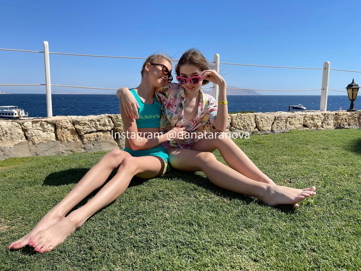 Two young women in sunglasses and summer outfits sit on green grass near the sea, posing together on a sunny day.