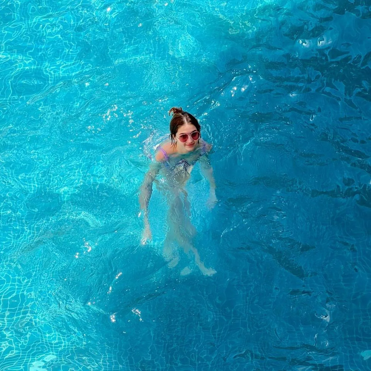 A woman with brown hair in a bun swims in clear blue pool water, viewed from above.