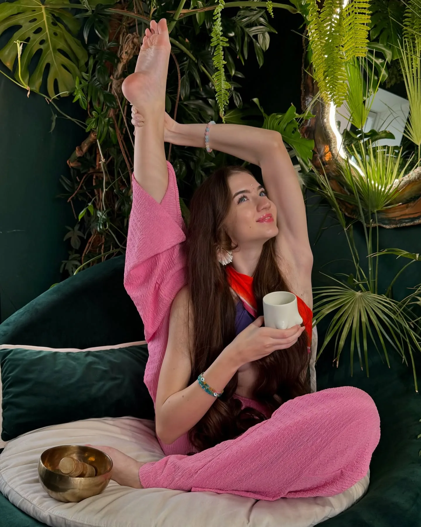 Woman in pink yoga pants performing a seated leg stretch while holding a mug, surrounded by lush green plants and a singing bowl.