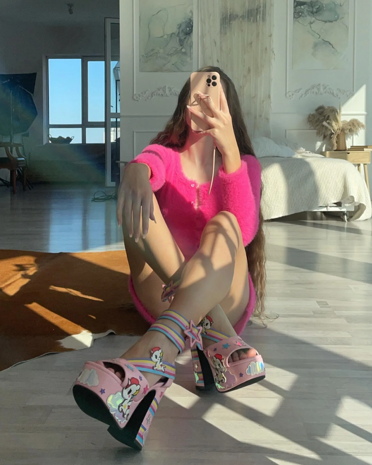 Woman in pink outfit and colorful platform heels taking a mirror selfie in a sunlit room.
