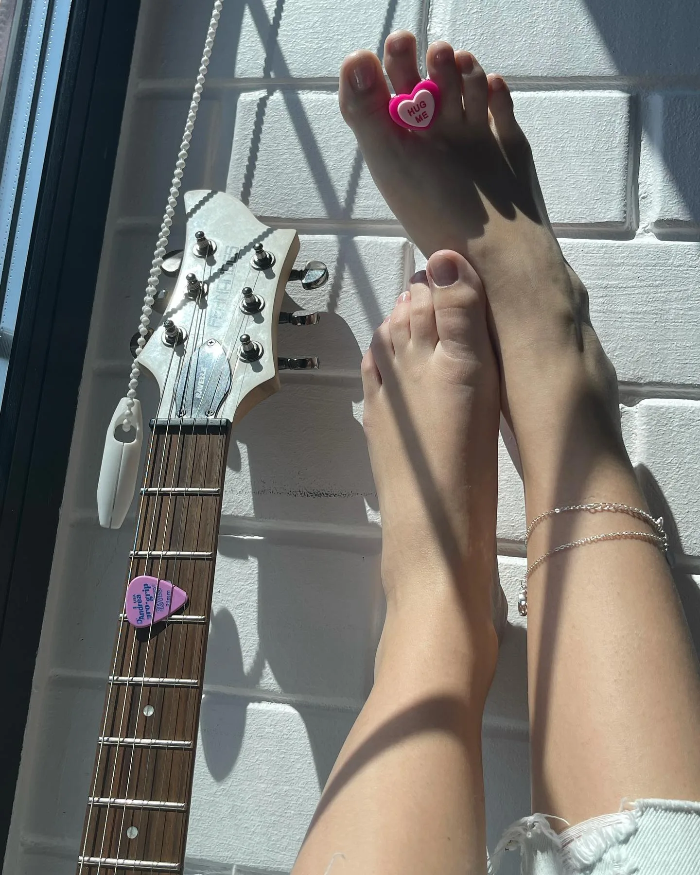 A pair of bare feet with a pink heart ring on the big toe and a delicate anklet, resting next to the headstock of a white electric guitar with a purple pick on the fretboard.