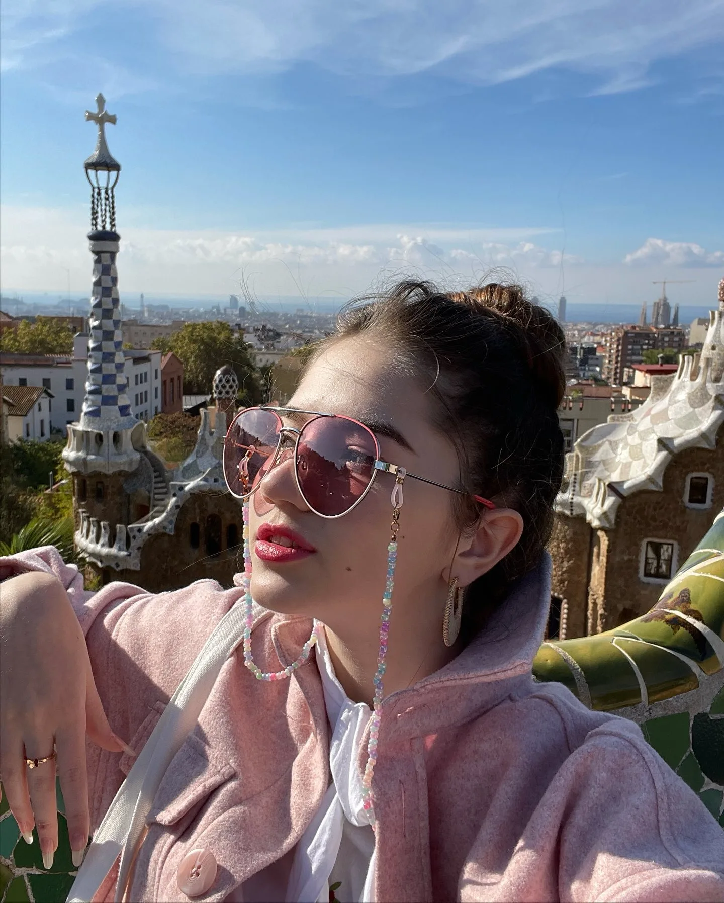 Dana wearing pink sunglasses and a pink jacket posing at Park Güell in Barcelona with the mosaic trencadís structures and city skyline in the background.