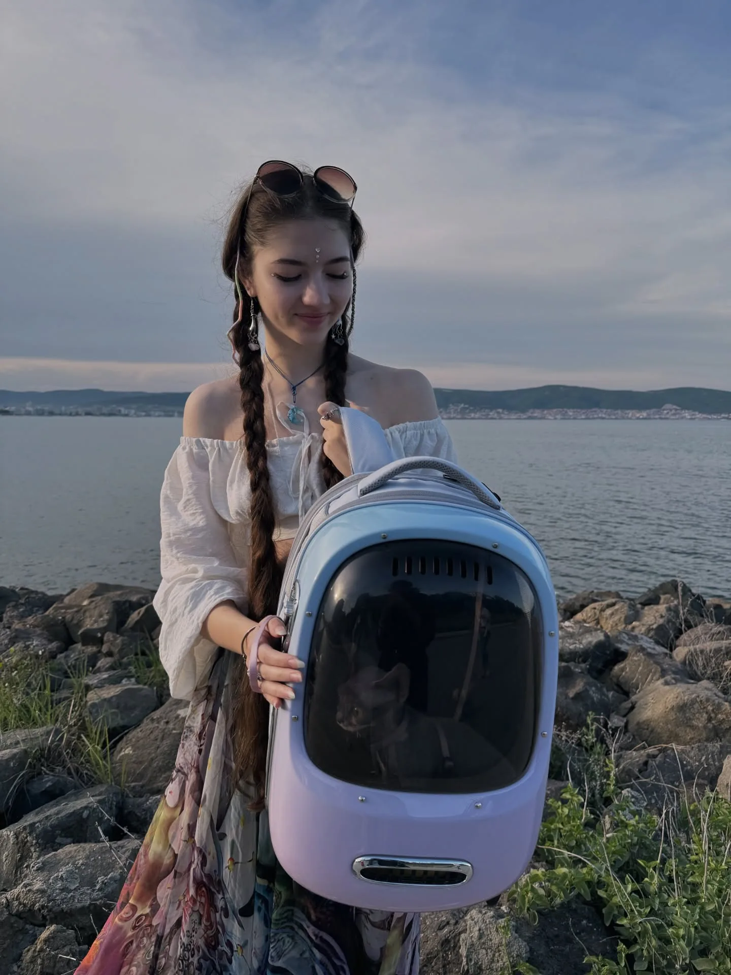 A woman with braided hair stands on rocky terrain near a body of water, holding a light purple and blue pet carrier backpack with a large transparent window.