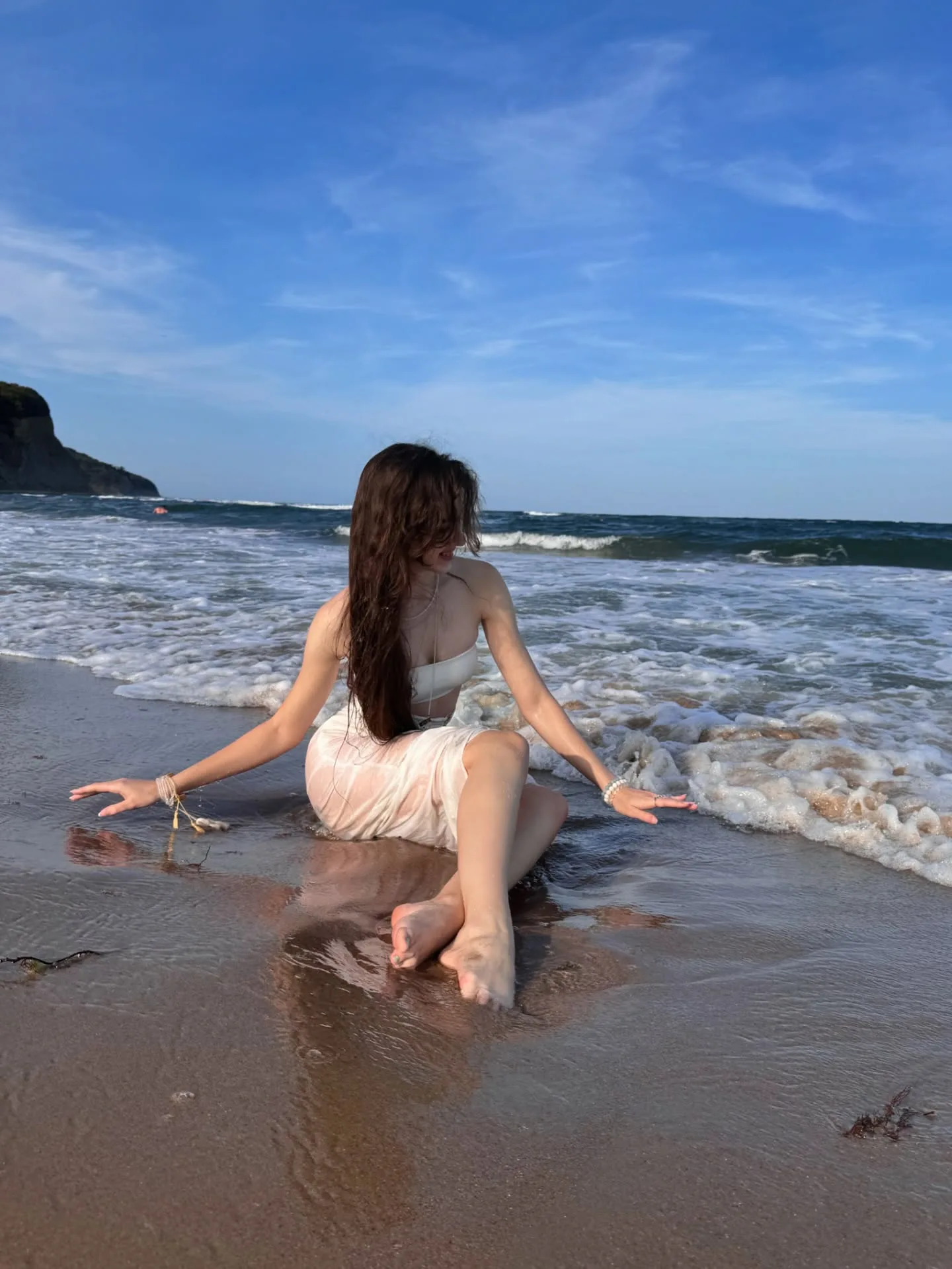 Woman in a white dress sitting on the wet sand at the beach with her back to the camera, looking out at the ocean waves and a distant headland under a blue sky.