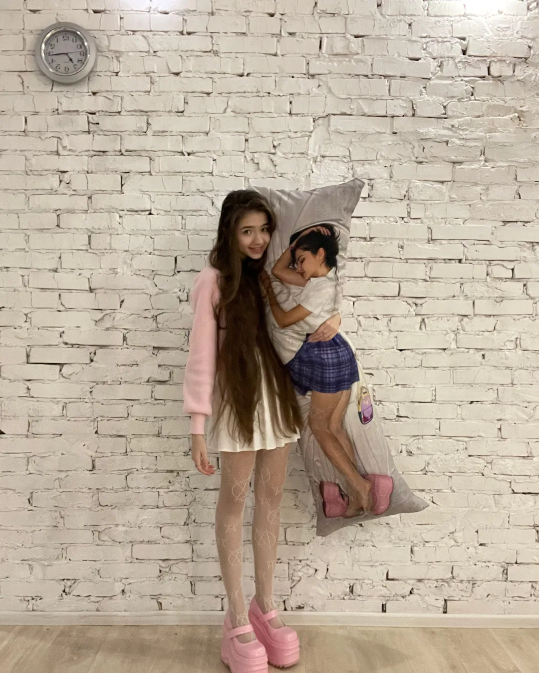 A young woman with long hair stands next to a large pillow featuring a printed image of a girl in a blue skirt and pink shoes against a white brick wall.