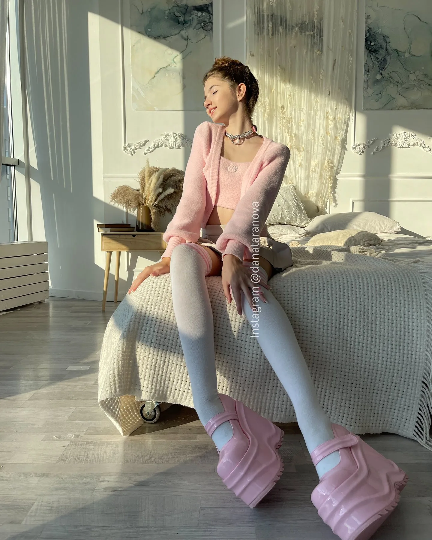 A woman in a pink cardigan and white tights sits on a bed wearing oversized pink platform shoes in a sunlit room.
