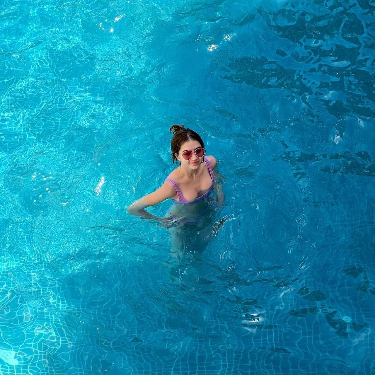 A woman with sunglasses and a pink swimsuit relaxes in a bright blue swimming pool, viewed from above.