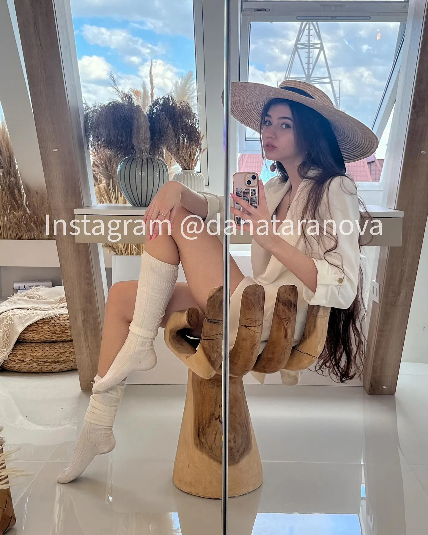Dana Taranova taking a mirror selfie in a wide-brimmed hat and white boots while seated on a wooden sculpture.