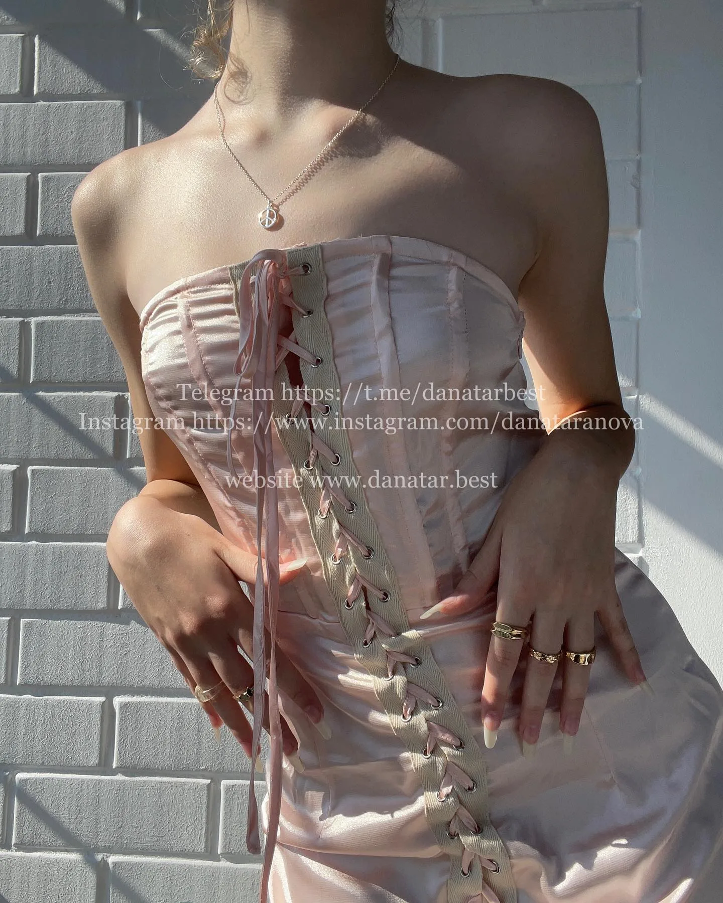 Dana wearing a pink satin corset dress with lace-up front, standing against a white brick wall.