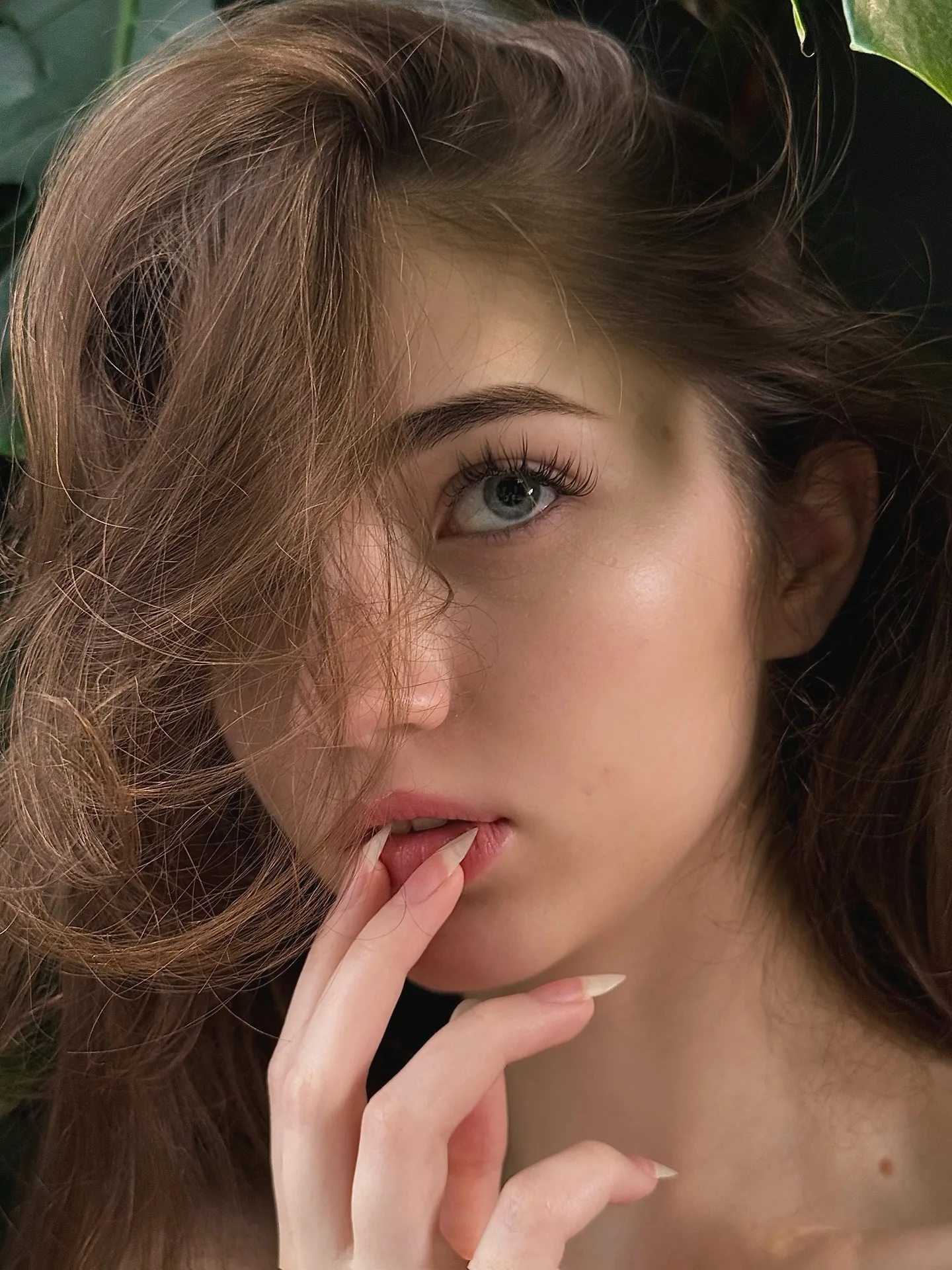 Close-up portrait of a young woman with brown hair and blue eyes, gently touching her lips with her fingers against a green leafy background.