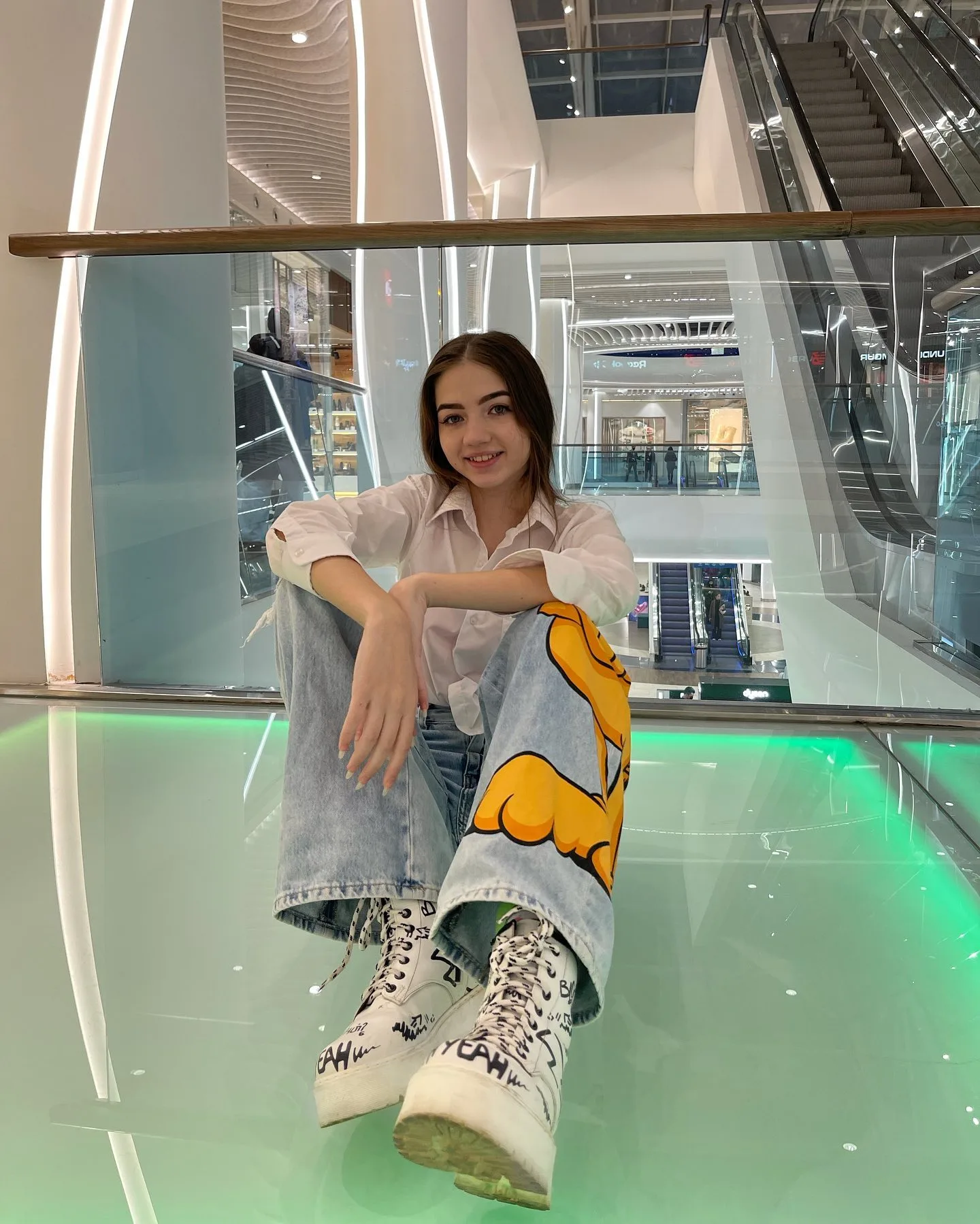 Dana sitting on a glass floor in a modern mall, wearing a white shirt, wide-leg jeans with a yellow graphic, and white platform boots.