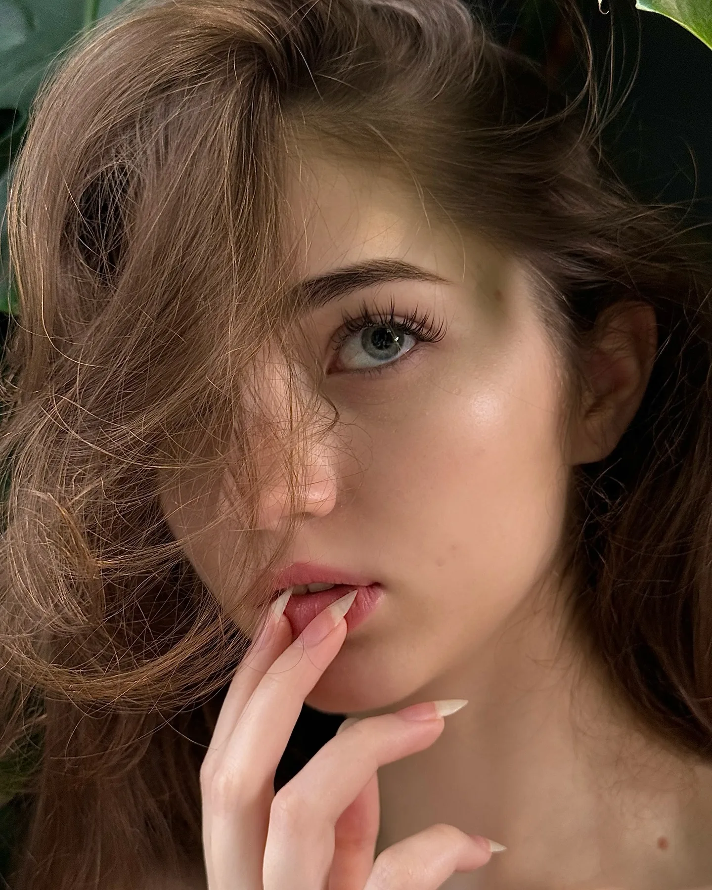 Close-up portrait of a young woman with wavy brown hair and blue eyes, gently touching her lips with her hand against a dark green background.