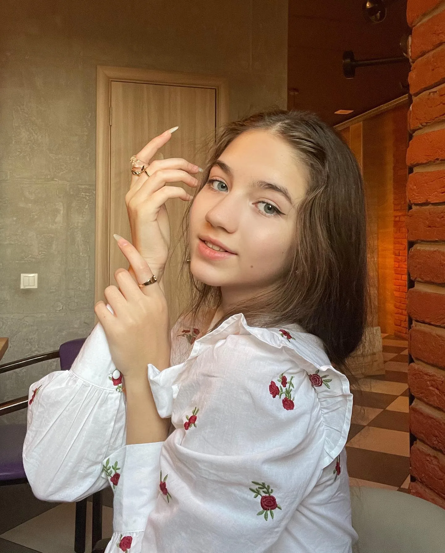 Portrait of Dana, a young woman with brown hair wearing a white blouse with red floral embroidery, posing indoors near a brick wall.