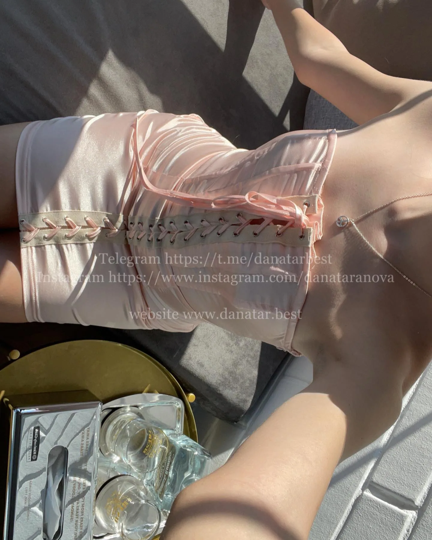 Close-up of a woman wearing a pink lace-up corset top and white shorts, with a gold tray and glasses in the foreground.