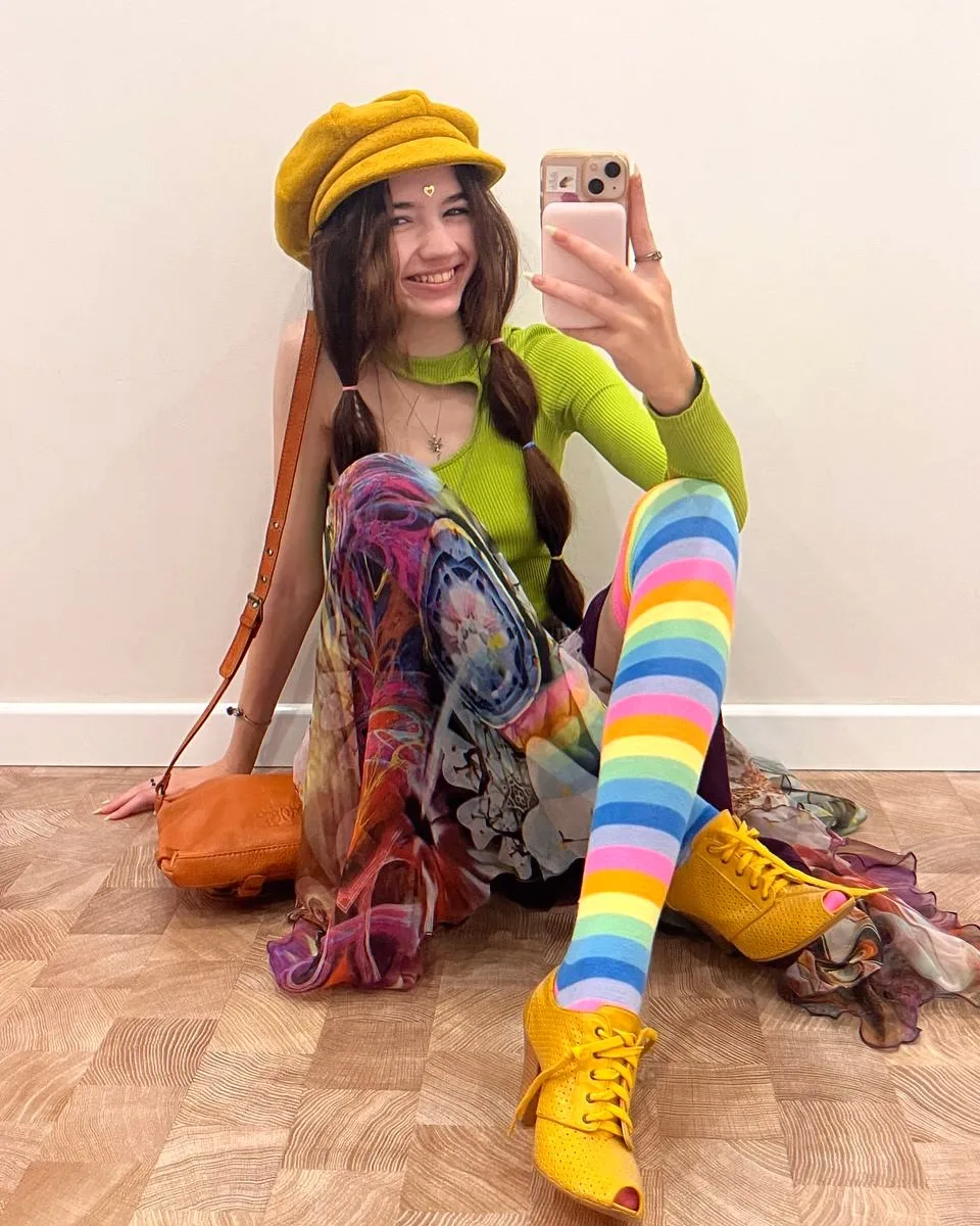 A smiling woman in a yellow beret, green top, and rainbow striped socks takes a mirror selfie while sitting on a wooden floor.