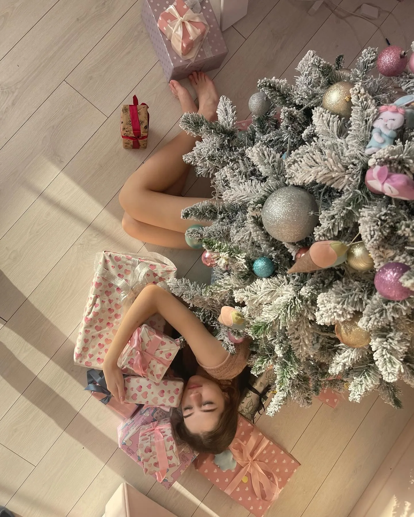A woman lying on a wooden floor next to a flocked Christmas tree and wrapped gifts.